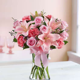 Pink lilies, roses, and carnations in a clear glass vase with a pink ribbon