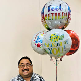 Smiling man holding three colorful get well message balloons in front of a beige wall