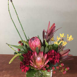 Mixed tropical bouquet in a clear square vase with pink protea and red blooms