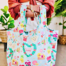 Pastel blue reusable tote bag with colorful cartoon flowers and characters held in front of a person.