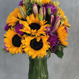 Bouquet of sunflowers, lilies, and purple flowers in a green glass vase