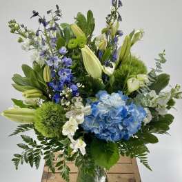 Tall arrangement of blue hydrangea, white lilies, and blue delphinium in a green glass vase