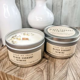 Two lidded tin candles labeled Black Currant + Jasmine on a wood block with white vases behind