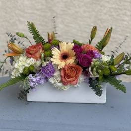 Mixed floral arrangement in a white rectangular vase with peach, purple, and white blooms