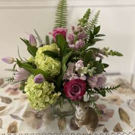 Spring arrangement with green hydrangeas, pink roses, and tulips in a glass vase beside a bunny figurine.