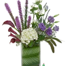 Tall mixed arrangement with white hydrangea, purple lisianthus, and calla lilies in a leaf-lined glass vase