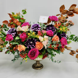 Wide arrangement of bright pink and orange roses with purple blooms and copper-toned branches in a pedestal vase