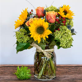 Bouquet of yellow sunflowers, peach roses, and green hydrangeas in a clear glass jar vase