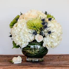 Low round arrangement of white hydrangeas, roses, and green mums in a leaf-wrapped glass bowl vase