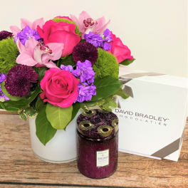 Pink and purple flower arrangement in a white vase with a candle and chocolate box