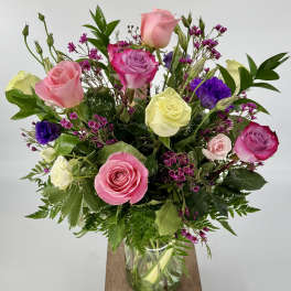 Mixed arrangement of pink, cream, and purple roses with small fuchsia blooms in a clear glass vase.