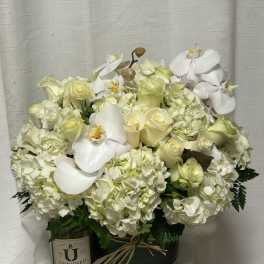 Low round arrangement of white roses, hydrangeas, and orchids in a green vase with a candle beside it