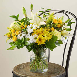 Yellow and white lilies, roses, and daisies arranged in a clear glass vase on a wooden chair