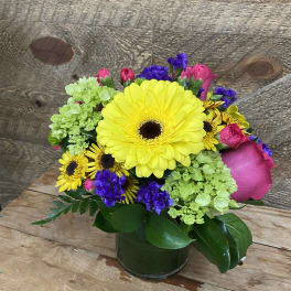 Mixed bouquet with yellow gerbera daisies, pink roses, and purple accents in a glass vase