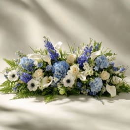 Low arrangement of blue hydrangeas, white calla lilies, roses, and anemones in a horizontal spray