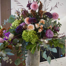 Large mixed arrangement of purple, pink, and green flowers with tall branches in a stone urn vase