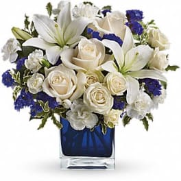 White lilies, roses, and carnations with blue accents in a dark blue glass cube vase