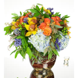 Low glass vase arrangement with blue hydrangeas, orange roses, yellow orchids, and purple blooms with trailing foliage.
