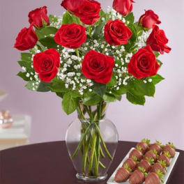 Red roses in a glass vase beside a tray of chocolate-covered strawberries