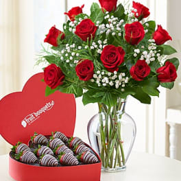 Red roses in a glass vase beside a heart-shaped box of chocolate-covered strawberries.