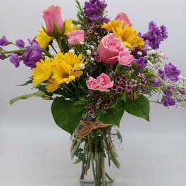 Mixed bouquet of pink roses, yellow daisies, and purple flowers in a glass vase