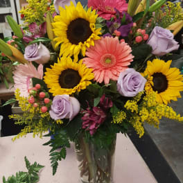 Mixed bouquet with sunflowers, gerbera daisies, and lavender roses in a glass vase