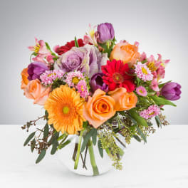 Bright mixed bouquet of orange roses, gerbera daisies, tulips, and pink blooms in a round clear glass vase