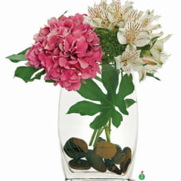Pink hydrangea and white alstroemeria in a clear glass vase with river rocks.