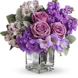Low arrangement of lavender roses, purple hydrangea, and mixed purple blooms in a clear glass cube vase