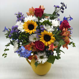 Mixed bouquet with roses, sunflowers, daisies, and blue flowers in a yellow pitcher