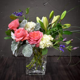 Arrangement of pink roses, white hydrangeas, and lilies in a clear glass cube vase with purple accents