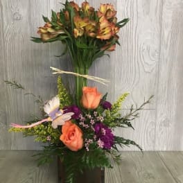 Tall mixed arrangement with orange roses, purple flowers, and alstroemeria in a brown glass cube vase