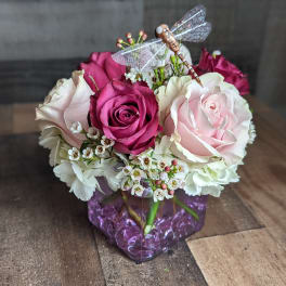 Short cube vase arrangement of pink and cream roses with white blooms and a decorative dragonfly pick.