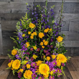 Tall one-sided arrangement of yellow roses and gerbera daisies with purple delphinium and daisy mums