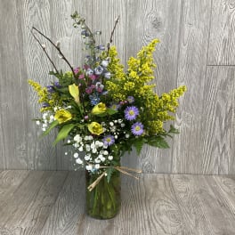 Tall mixed bouquet in a glass vase with yellow, purple, white, and pink flowers.