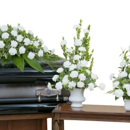 White carnation funeral casket spray with two matching vased arrangements beside a dark casket