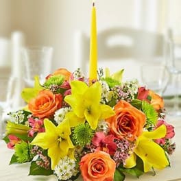 Low floral centerpiece with yellow lilies, orange roses, and a tall yellow candle