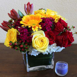 Colorful bouquet of roses and daisies in a square glass vase beside a blue candle