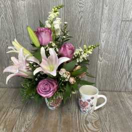 Pink roses and lilies arranged in a floral mug with a matching mug beside it.
