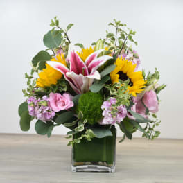 Bright mixed arrangement with sunflowers, pink lilies, and pastel blooms in a square glass vase