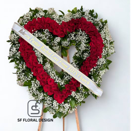 Heart-shaped standing wreath of red roses and white filler flowers with a printed ribbon across the front.