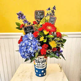 Bright mixed flower arrangement in a tall teacher-themed vase with chalkboard picks.