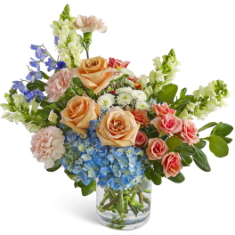 Mixed bouquet of peach roses, blue hydrangea, and assorted blooms in a clear glass vase