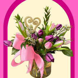 Pink and purple tulip bouquet in a glass vase with a ribbon and heart topper