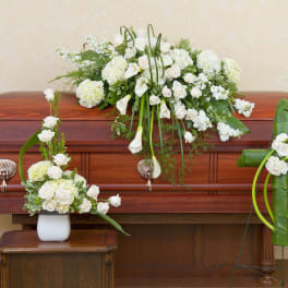 White funeral spray, vase bouquet, and cross arrangement displayed on a wooden casket