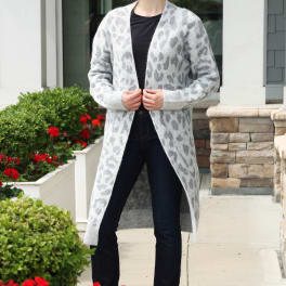 Woman in a long gray patterned cardigan and dark jeans standing by white planters with red flowers