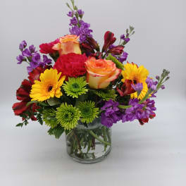 Mixed bouquet of roses, gerbera daisies, carnations, and purple blooms in a glass vase