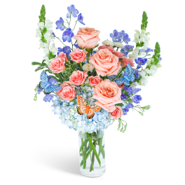 Peach roses and blue flowers in a clear glass vase with butterfly accents