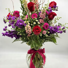 Mixed bouquet of red roses, purple blooms, and pink flowers in a glass vase with ribbon
