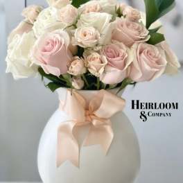 Pink and white roses arranged in a white vase with a peach ribbon